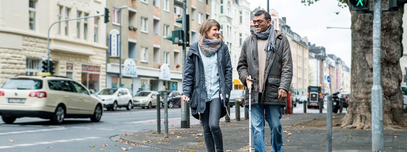 Frau und Mann mit Blindenstock gehen über eine Straße
